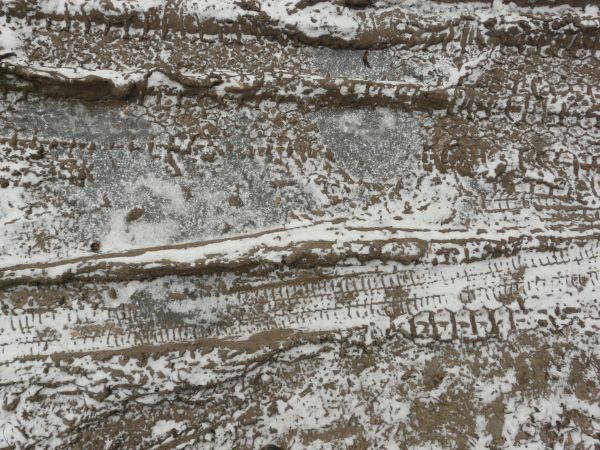 Dirt road texture in dark brown tone with tire marks and thin layer of snow across surface.